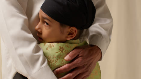 Nahaufnahme-Studioaufnahme-Eines-Jungen-Sikh-Jungen-Mit-Turban-Und-Haarknoten,-Der-Seinen-Großvater-Vor-Einfarbigem-Hintergrund-Umarmt-2